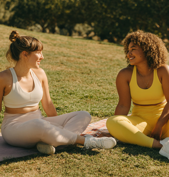 Two women in activewear sitting and talking on the grass in a park